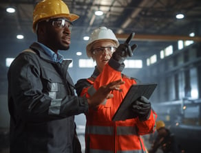 Man and Woman in hardhats pointing in dark factory - shutterstock_1870469602 (1) (1)